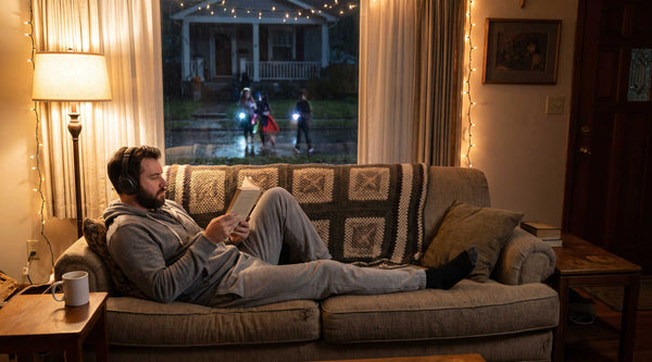 Man in headphones reading on the couch on Halloween night while trick-or-treaters pass outside the window