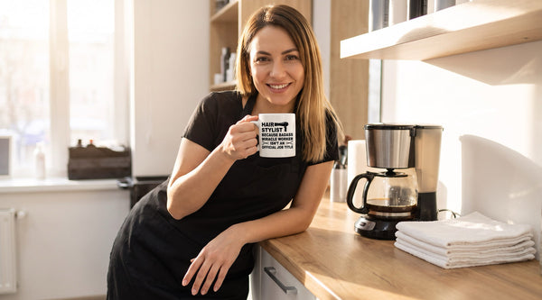 Hair stylist in a black apron laughing on a coffee break in the salon break room, holding a funny hair stylist coffee mug