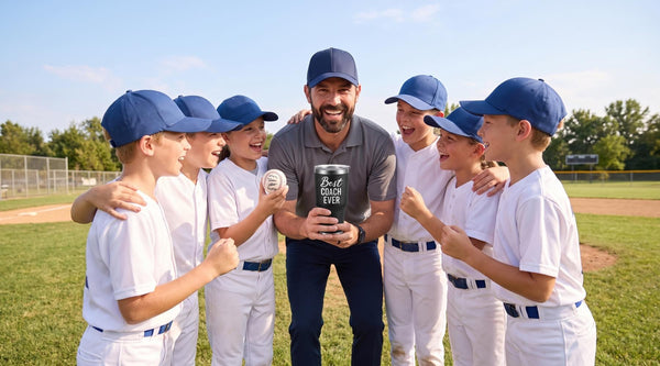 A Little League team in white uniforms cheers around their coach on the field as he holds a Best Coach Ever tumbler and a player holds a team-signed baseball