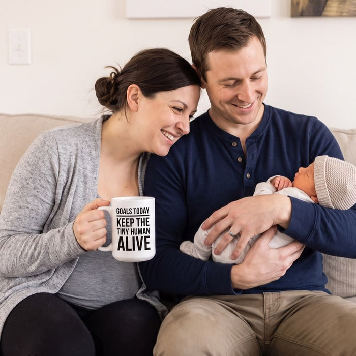 Working mom smiling on sofa holding Goals Today Keep The Tiny Human Alive mug with partner and newborn baby