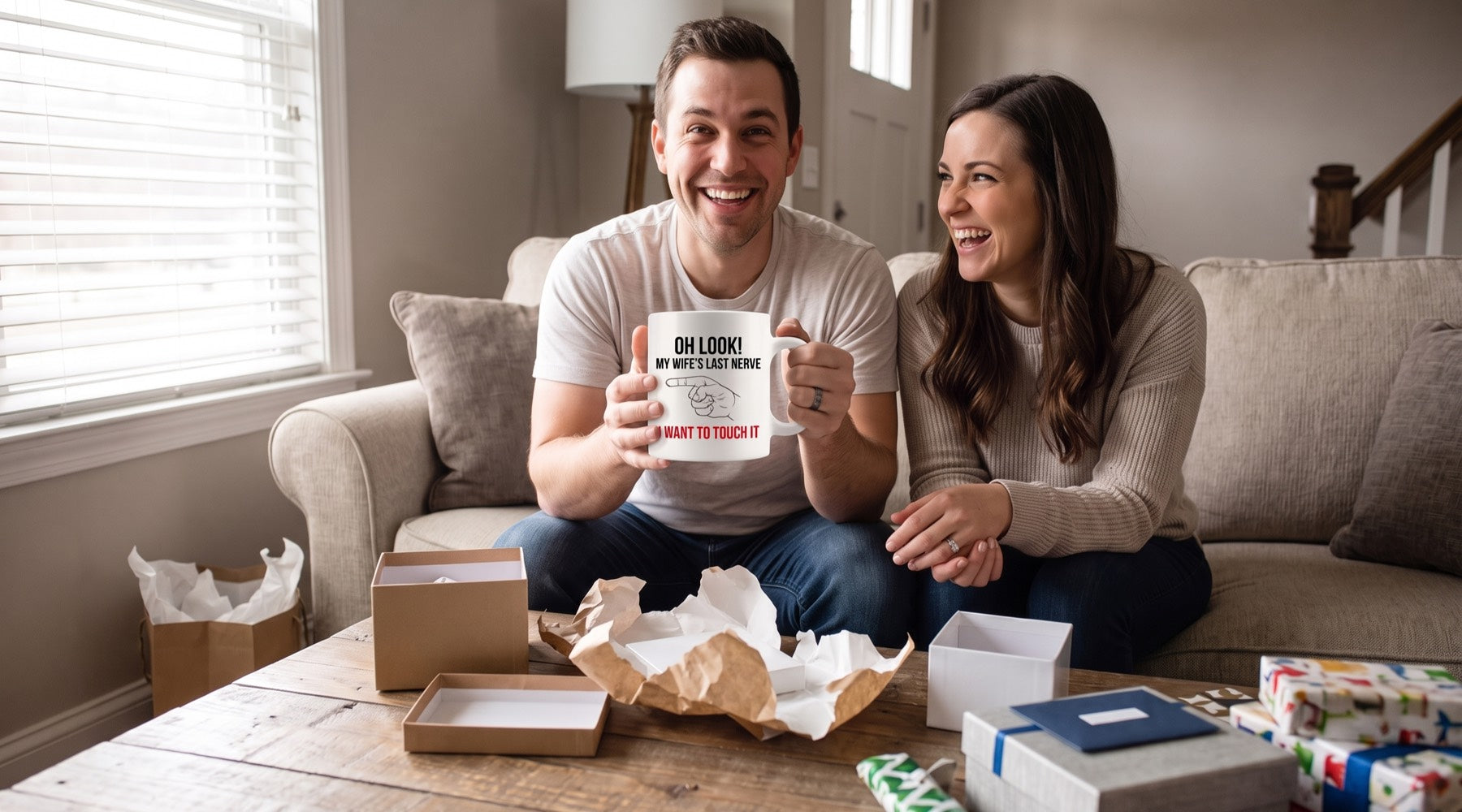 Newlyweds laugh on their sofa among opened wedding gifts as the husband holds an Oh Look My Wife's Last Nerve mug