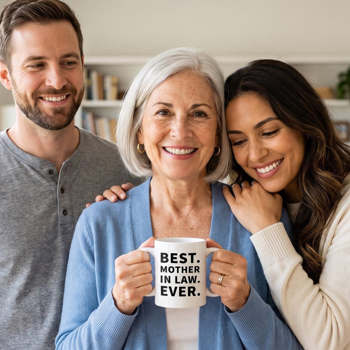 A mother-in-law holds a Best Mother In Law Ever mug while her daughter-in-law hugs her and her son stands beside them in a bright living room