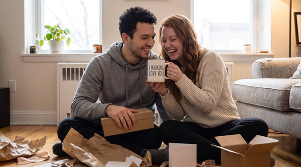 Young newlywed couple laughing on their living room floor as she unwraps an I Freaking Love You mug among wedding gifts