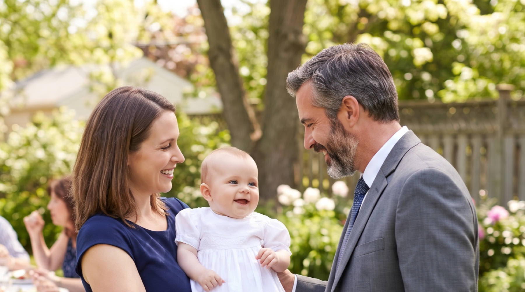 A godmother holding a smiling baby in a white baptism gown while the godfather smiles at the baby in a garden